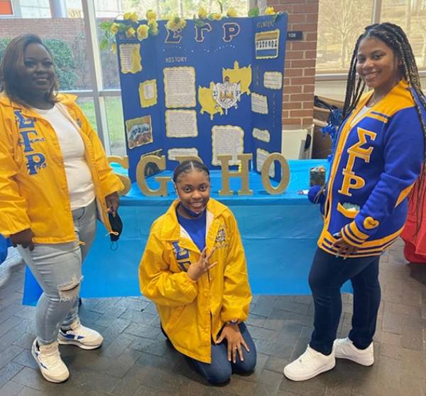 Image of members of Rho Lambda Chapter of Sigma Gamma Rho standing in front of their tri-fold board, wearing blue and gold Greek Letter paraphenalia including windbreaker jacketes and a fleece coat.