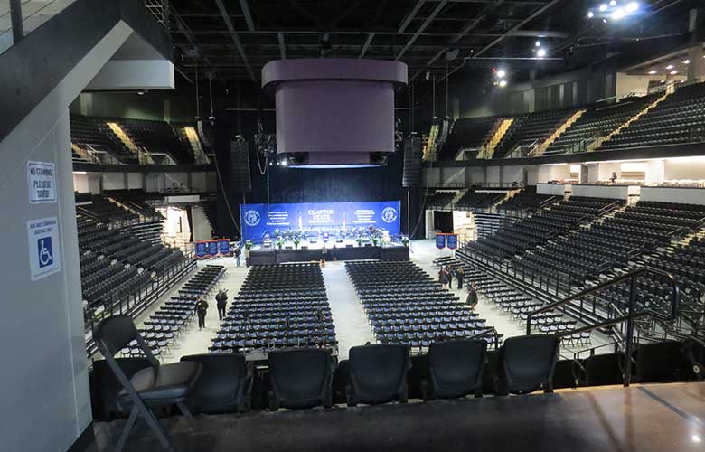 Interior view of a large arena set up for a commencement ceremony with rows of floor seating facing a stage. Tiered accessible seating surrounds the venue, with wide aisles, railings, and wheelchair-accessible areas visible throughout the spacious event space.