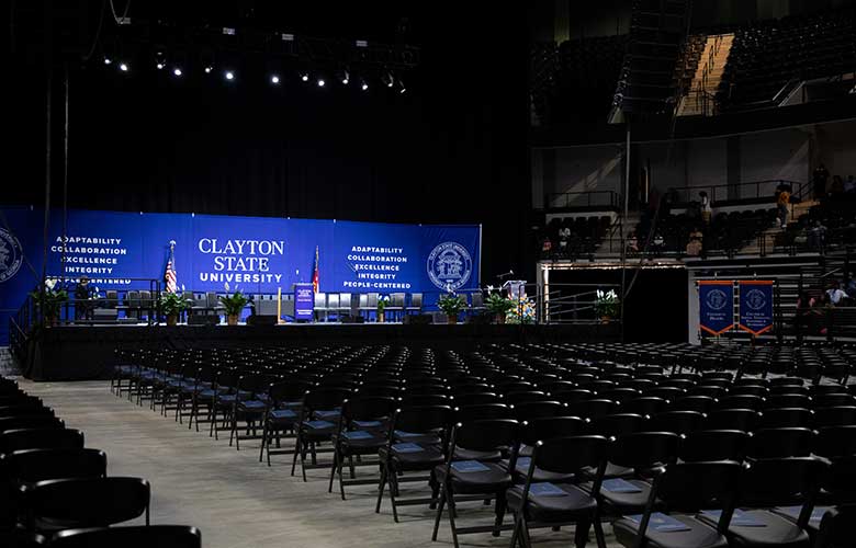 Rows of chairs fill an arena floor leading to a commencement stage decorated with Clayton State University banners, flags, and ceremonial furnishings. The venue is prepared for guest entry and seating, with clear aisles and designated sections surrounding the stage.