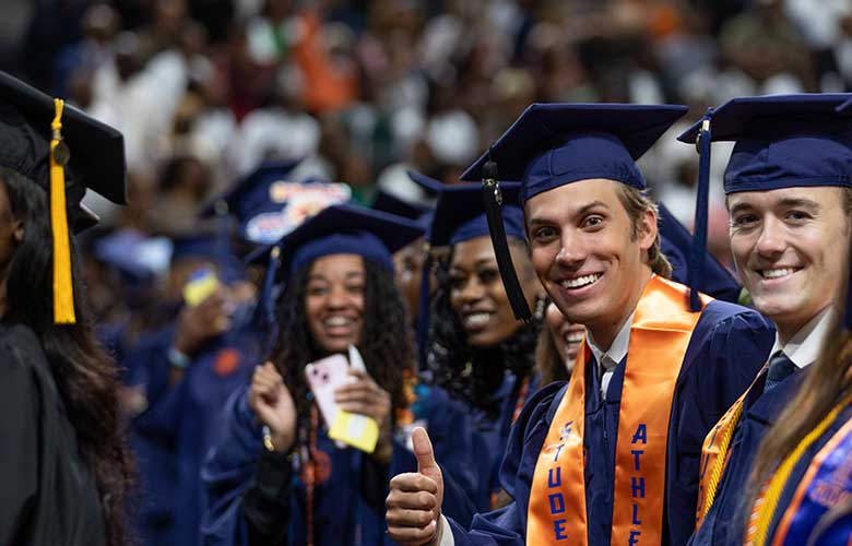 Graduates in blue caps and gowns sit among fellow classmates during a commencement ceremony. One graduate in the foreground gives a thumbs-up while wearing an orange honor stole. A crowded arena audience fills the background, celebrating the academic milestone.