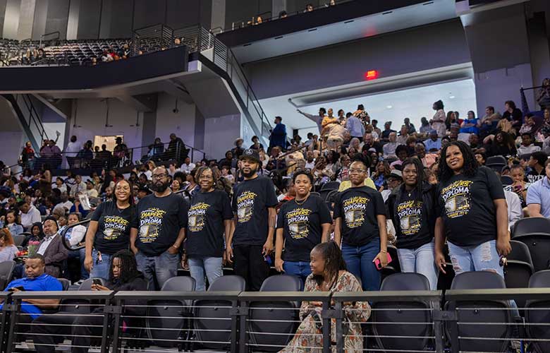 Guests gather in arena seating during a commencement ceremony. A group wearing matching celebratory shirts stands together in the foreground while attendees fill surrounding sections. The venue is busy with family and friends gathered to support graduating students.