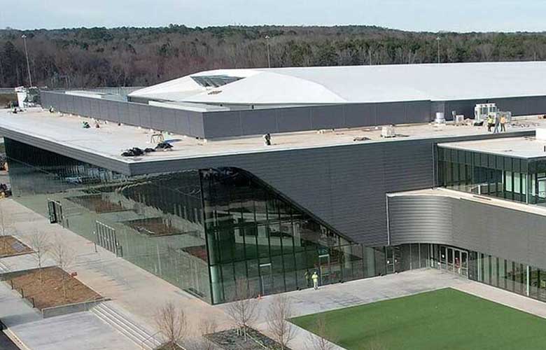 Exterior view of a modern arena and event venue with expansive glass walls, curved architectural features, and surrounding walkways. The facility offers guest parking and access points for commencement attendees, with landscaped grounds and entrances visible around the building.