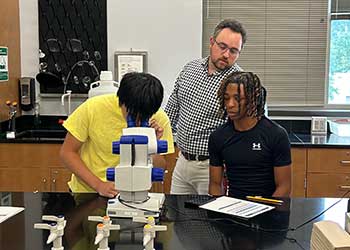 Students conduct hands-on biology research in a science laboratory while examining a specimen under a microscope. An instructor provides guidance as students observe and record findings at a lab workstation equipped with scientific tools and materials.