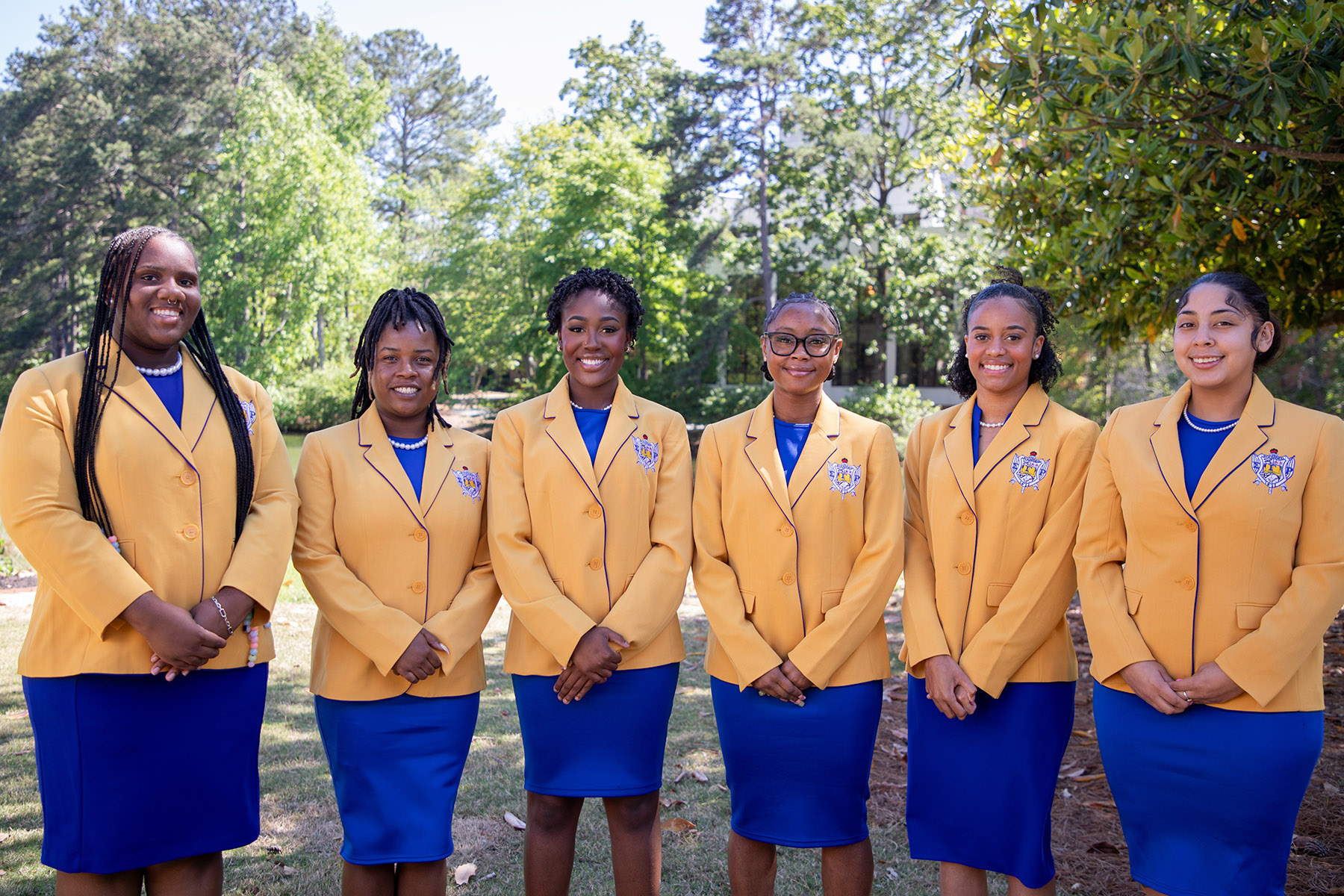 Image of members of Rho Lambda Chapter of Sigma Gamma Rho standing in a line, facing the camera, wearing gold jackets with their seal on the pocket, blue dresses, and a string of pearls.