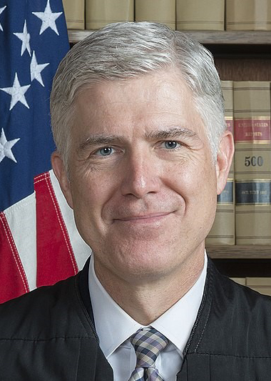 A portrait of Justice Neil Gorsuch, wearing a black judicial robe, smiling slightly. His short, gray hair is neatly combed, and he is positioned in front of a background of law books, indicating a judicial or legal setting.